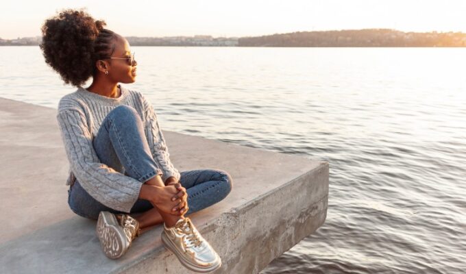 Black woman sitting close to water at a beach practicing daily calm