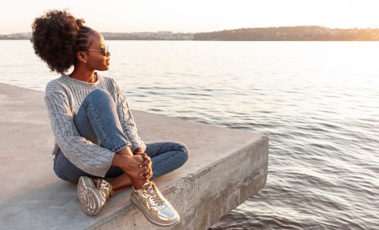 Black woman sitting close to water at a beach practicing daily calm