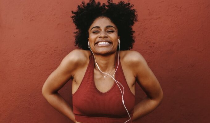 A Black woman with natural hair wearing headphones and red sports bra excited about fitness for beginners.