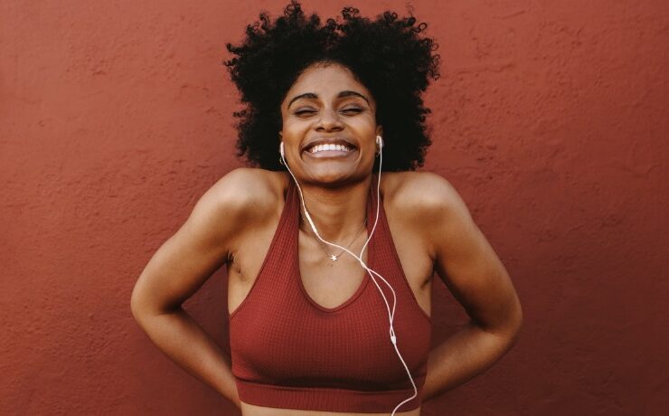 A Black woman with natural hair wearing headphones and red sports bra excited about fitness for beginners.
