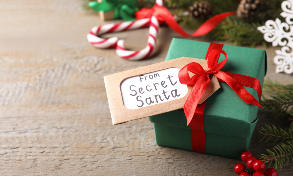 Small green Secret Santa gift box with red ribbon and tag on a rustic wood surface, surrounded by festive Christmas decorations.