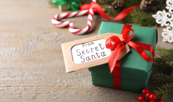 Small green Secret Santa gift box with red ribbon and tag on a rustic wood surface, surrounded by festive Christmas decorations.