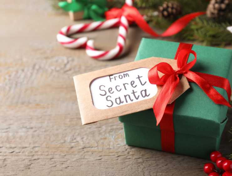 Small green Secret Santa gift box with red ribbon and tag on a rustic wood surface, surrounded by festive Christmas decorations.